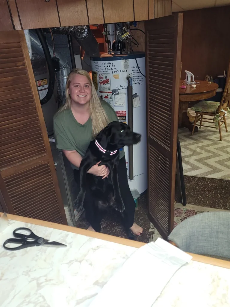 Brittany Alms kneeling with her black Labrador in front of her old water heater
